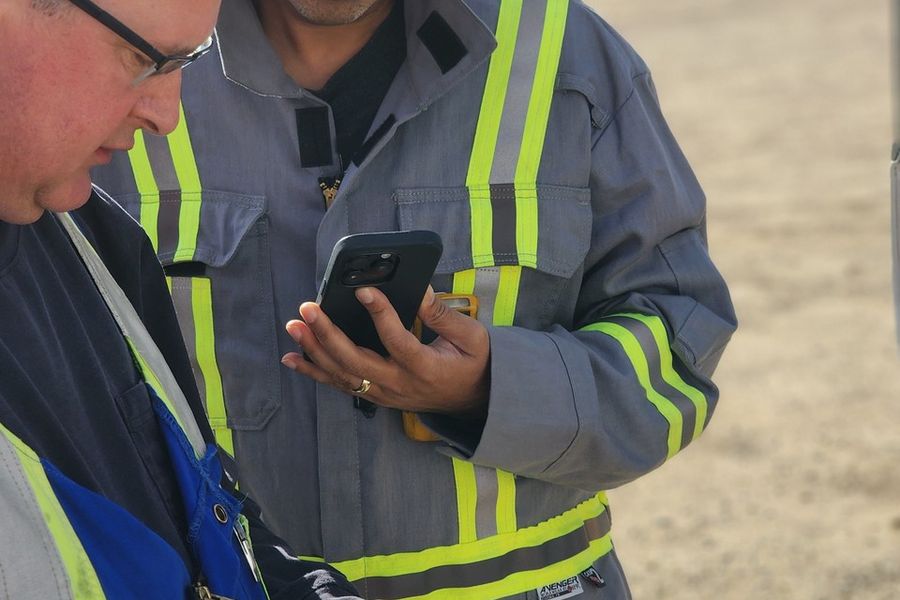 Two field workers in hi-vis coveralls at a facility, one checking a phone and the other working on a rugged tablet.